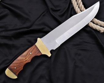 May include: A large, stainless steel knife with a polished blade and a wooden handle. The handle is a dark brown color with brass accents. The knife is displayed on a black surface with a rope in the background.