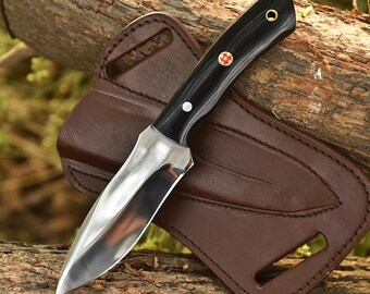 May include: A handcrafted hunting knife with a polished steel blade and a black wooden handle. The knife rests on a brown leather sheath, showcasing a classic design. The handle features a decorative red and white emblem.