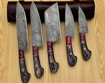 May include: Set of five kitchen knives with Damascus steel blades and wooden handles. The knives have a red and black pattern on the handles. The knives are arranged in a row on a wooden surface.