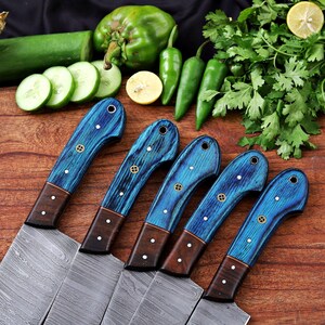 May include: Five Damascus steel chef knives with blue wooden handles and brown accents are arranged on a wooden surface. The handles have a unique grain pattern and are adorned with decorative rivets. Fresh vegetables, including cucumbers, peppers, and lemons, are in the background.