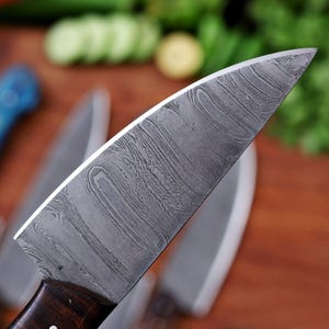 May include: Close-up of a Damascus steel chef's knife with a dark wooden handle. The blade has a distinctive wavy pattern. Other knives are slightly visible in the background, along with a wooden cutting board and some vegetables.