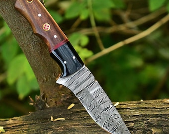 May include: A Damascus steel knife with a wooden handle. The handle is a dark brown color with brass rivets and a black section near the blade. The blade has a unique swirling pattern. The knife is resting on a tree branch.