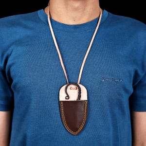 May include: A leather pendant necklace with a brown, shield-shaped pouch and a cream-colored top section. The pendant is suspended from a thin, light-colored leather cord. The pouch has yellow stitching around the edges.
