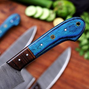 May include: Close-up of a set of kitchen knives with blue and brown wooden handles. The handles have a vibrant blue wood grain pattern with decorative metal accents. The blades are silver and the knives are arranged on a wooden surface.