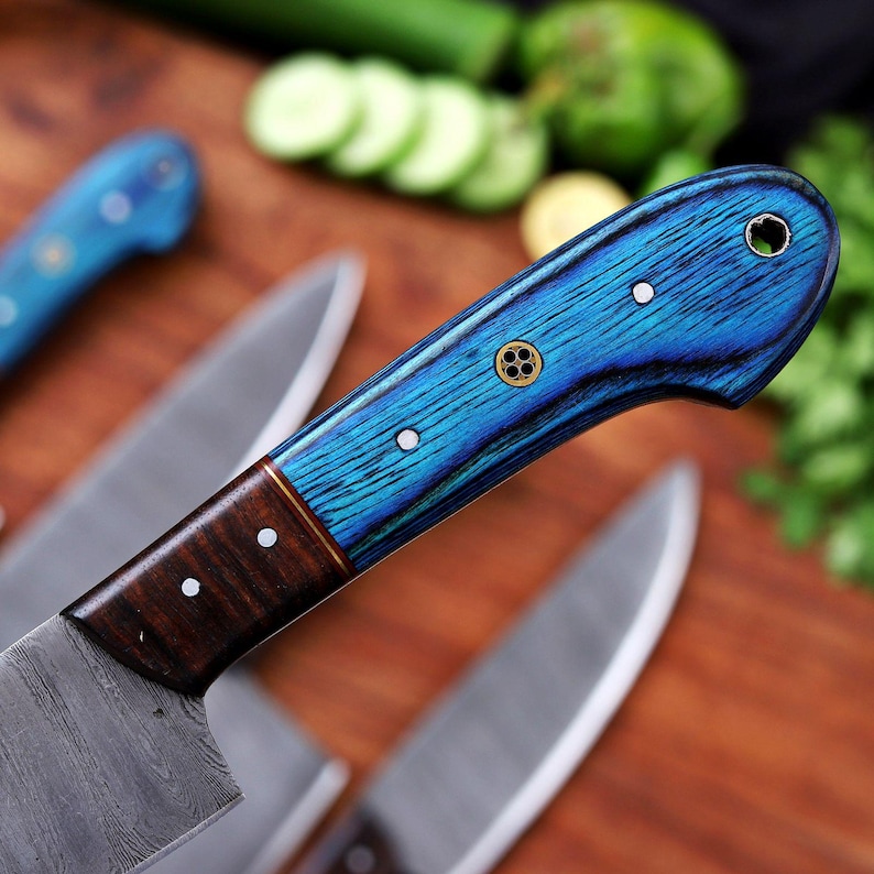 May include: Close-up of a set of kitchen knives with blue and brown wooden handles. The handles have a vibrant blue wood grain pattern with decorative metal accents. The blades are silver and the knives are arranged on a wooden surface.