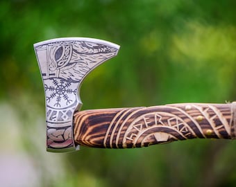 May include: Close-up of a silver axe head with etched Viking-inspired designs, including runes and a snowflake symbol. The wooden handle features carved patterns and is wrapped with twine. The axe is set against a blurred green background.