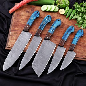 May include: A set of six kitchen knives with patterned steel blades and blue wooden handles. The handles have a dark brown bolster and are secured with small brass rivets. The knives are arranged on a wooden cutting board.
