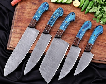 May include: A set of six kitchen knives with patterned steel blades and blue wooden handles. The handles have a dark brown bolster and are secured with small brass rivets. The knives are arranged on a wooden cutting board.