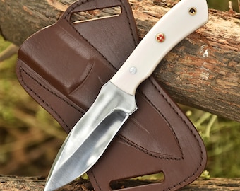 May include: A hunting knife with a polished steel blade and a cream-colored handle, accented with brass rivets and a red and gold emblem. The knife rests in a brown leather sheath with a belt loop.