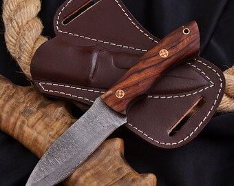 May include: A Damascus steel knife with a wooden handle and brass rivets rests in a brown leather sheath. The knife blade has a unique pattern. The sheath is stitched with white thread. A rope is in the background.