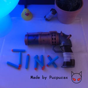 以下が含まれることがあります： メタリック仕上げの小道具銃。詳細なデザインで、青色の「JINX」の文字が特徴です。青とオレンジのおもちゃのダーツが含まれています。「Made by Purpurax」の文字も表示されています。