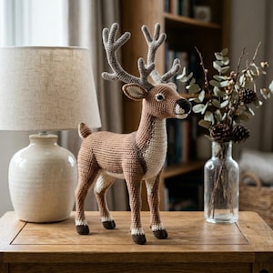 May include: A crocheted deer ornament in brown and cream tones, with detailed antlers and hooves. The deer is displayed on a wooden table, alongside a lamp and a vase with branches and pine cones. A decorative item for the home.