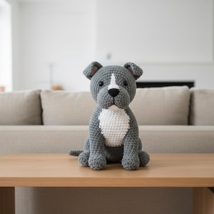 May include: A crocheted dog toy in grey with white markings on its chest and face. The dog is sitting on a light brown wooden table. The toy has a black nose and small, dark eyes. The table is in front of a beige sofa.