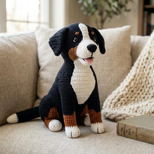 Pode incluir: Um cão de crochê feito à mão em posição sentada. O cão tem cores preto, branco e marrom, com um peito branco e uma faixa branca na cabeça. O cão está sentado em um sofá bege.