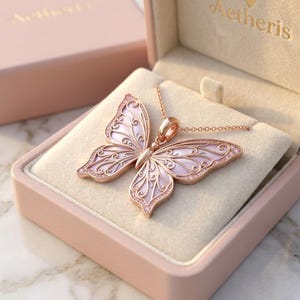 May include: A rose gold butterfly pendant necklace featuring iridescent pink inlay and intricate filigree metalwork. The pendant rests inside a light pink jewelry box with the brand name Aetheris embossed in gold on the interior lid.