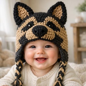 May include: A smiling baby wearing a handmade crochet hat shaped like a German Shepherd dog. The hat is black and tan, with ear flaps and braided tassels. The baby is wearing a cream-colored onesie.