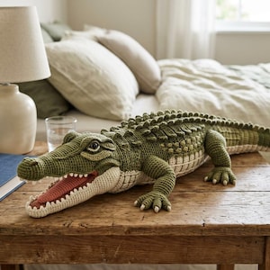May include: A large, handmade crochet crocodile in shades of green and cream, with an open mouth revealing a red interior. The plush toy is displayed on a wooden table, showcasing its detailed craftsmanship and playful design.