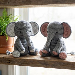May include: Two gray crochet elephant stuffed animals with pink and white accents sit on a wooden shelf. The elephants have large ears, trunks, and small tails. A fern and a ceramic jar are in the background.