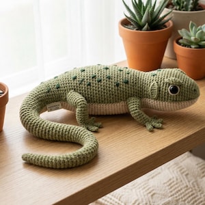 May include: A handmade, green crochet lizard with dark green spots, lying on a wooden surface. The lizard has a cream-coloured belly and black eyes. The tag reads "HANDMADE BY LUNA". Several potted plants are in the background.
