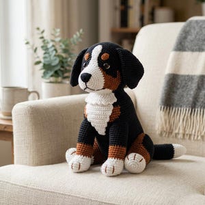 Puede incluir: Un peluche hecho a mano de un perro de montaña bernés tejido a ganchillo. El perro es negro, marrón y blanco, con el pecho y las patas blancas. Está sentado en un sillón crema, con una manta y una planta en el fondo.