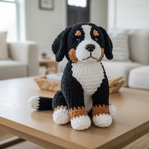 Peut inclure: Une peluche faite à la main représentant un chien de montagne bernois au crochet. Le chien a des marques noires, blanches et marron, avec une poitrine et des pattes blanches. Il est assis sur une table en bois brun clair.