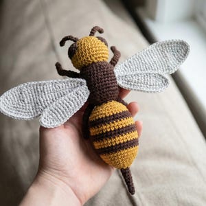 May include: A handmade crochet bee with a yellow and brown striped body, brown head, and gray wings. The bee is a soft sculpture, perfect for a child's toy or nursery decor. The bee is approximately 6 inches long.