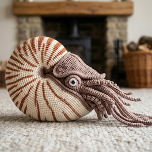 Op de afbeelding: Een gehaakte nautilus sculptuur in crème en bruin. De schelp heeft bruine strepen en de kop heeft gedetailleerde tentakels. De sculptuur staat op een neutraal tapijt. Een decoratief item voor in huis.