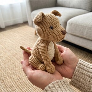 May include: A crocheted dog toy in shades of tan and cream. The handmade puppy has a brown nose and eyes, with a cream-colored patch on its chest. The dog is sitting, with its tail curled, and is held in cupped hands.