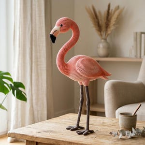 Könnte beinhalten: Ein handgefertigter, rosa gehäkelter Flamingo mit langem Hals und Beinen. Der Flamingo hat einen schwarzen Schnabel und steht auf einem Holztisch. Der dekorative Vogel ist eine charmante Ergänzung für jede Wohnkultur.