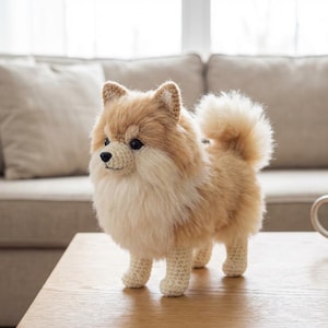 以下が含まれることがあります： 手作りの、かぎ針編みのポメラニアン犬の置物。犬はふわふわの薄茶色とクリーム色の毛並みで、顔と足はかぎ針編みで細かく作られています。木製のテーブルの上に立っており、背景にはニュートラルカラーのソファとマグカップがあります。