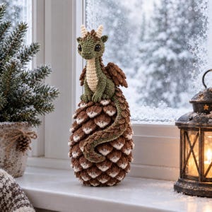 May include: A crocheted green dragon figurine with cream-colored horns and belly, perched atop a large, textured brown and white pinecone. The dragon has dark, round eyes, small wings, and a long, winding tail that curls down the pinecone surface.