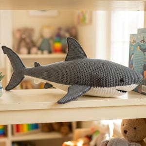 May include: A handmade crochet shark plush toy featuring a dark gray top and a white underside. The shark has a prominent dorsal fin, side fins, a tail, and small black bead eyes. The mouth is slightly open, revealing small white crochet teeth.