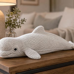 May include: A light gray crocheted dolphin-shaped pillow with black eye details. The plush toy features fins and a tail, resting on a wooden surface. The dolphin is a decorative item for a nursery or living room.