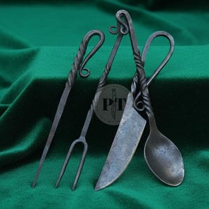 May include: Four hand-forged iron kitchen utensils: a carving fork, a knife, a spoon, and a serving fork. Each tool has a twisted handle and a dark, rustic finish. The tools are arranged against a green velvet backdrop.