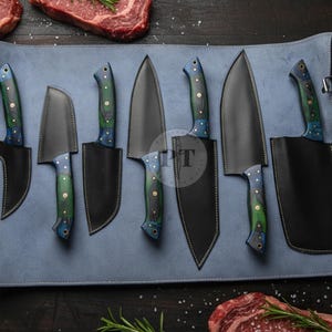 Può includere: Un set di sette coltelli da cucina con lame nere e manici con motivi a venature blu e verdi. I coltelli sono esposti su un rotolo di pelle blu con sfondo in legno scuro e bistecche crude.