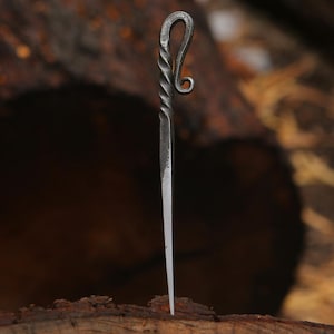 May include: A hand-forged metal hair pin featuring a dark grey finish, a twisted shaft design, and a decorative curled top loop. The pointed end tapers to a fine tip, showcasing a textured, industrial aesthetic with visible metalwork details.