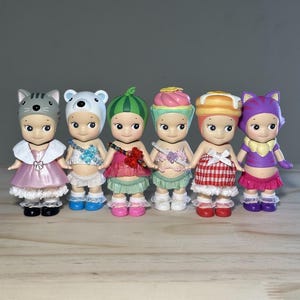 May include: Six small figurines with unique outfits and headwear. The figurines include a cat, bear, watermelon, cupcake, donut, and cat-themed designs. Each figure has a distinct outfit, including dresses, skirts, and tops in various colors like pink, red, and purple.