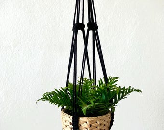 May include: Black macrame plant hanger with a woven basket and a green plant inside. The hanger has a tassel at the bottom.