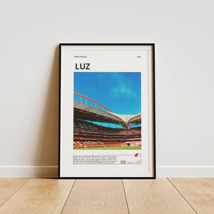 Może przedstawiać: Czarny plakat w ramie z tekstem "LUZ" w białych literach. Plakat przedstawia zdjęcie dużego stadionu z błękitnym niebem i białymi chmurami. Stadion ma czerwono-białą kolorystykę. Tekst "Lizbona, Portugalia" znajduje się w lewym górnym rogu plakatu.