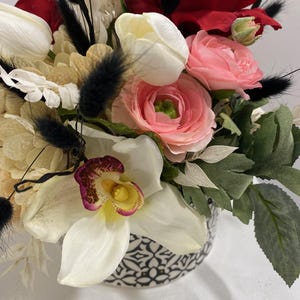 May include: A white ceramic vase with a black and white geometric pattern is filled with a bouquet of artificial flowers. The bouquet includes white orchids, pink and red roses, white tulips, and white and pink ranunculus.