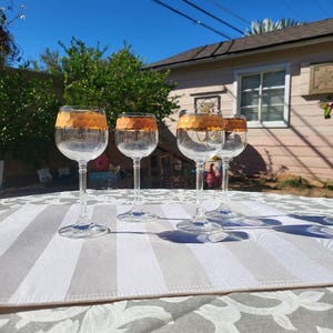 May include: Four clear wine glasses with a gold honeycomb design around the rim are arranged on a striped tablecloth. The glasses have long stems and sit on a table outdoors, with a house and trees in the background.