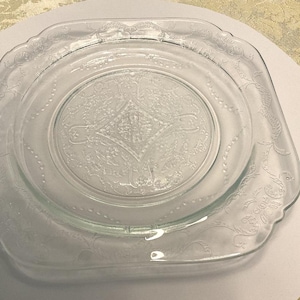 May include: Clear glass serving dish with a square shape and a decorative floral and geometric pattern. The dish has a raised rim and a circular indentation in the center. The glass is transparent, allowing light to pass through.