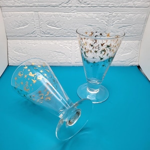 May include: Two clear, stemmed glasses with a flared top and a round base. The glasses are decorated with gold and green celestial designs. One glass is upright, and the other is lying on its side. The background is a white brick wall and a blue surface.