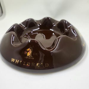 May include: A glossy, brown ashtray with a wavy rim. The ashtray features the text "WHITBREAD" in gold lettering, along with a logo and the year "1742". The ashtray is a vintage item.