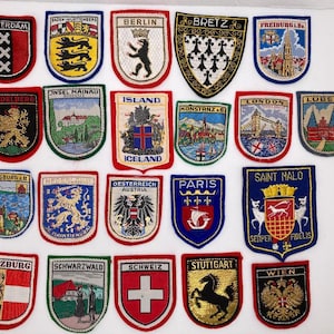 May include: A collection of twenty-two embroidered souvenir patches featuring various European city and country emblems. Each patch displays distinct colorful designs, including heraldic animals, architectural landmarks, and regional coats of arms.
