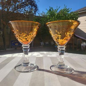 May include: Two amber-colored, stemmed glasses with textured bowls. The glasses have a clear stem and base, and are set on a striped surface. The glasses are likely vintage or antique, and are suitable for serving cocktails or other beverages.