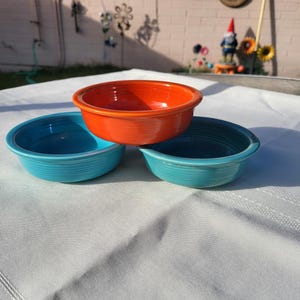 May include: Three colorful ceramic bowls in shades of orange and turquoise. The bowls are stacked on a white tablecloth, with the orange bowl on top. The bowls have a classic, simple design.