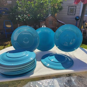 May include: A collection of turquoise-colored ceramic plates and saucers. The plates are round with a concentric ring design, and the saucers have a raised center. Some plates are stacked, while others are displayed on clear stands. The setting is outdoors.