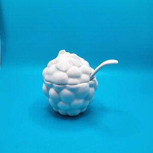 May include: A white ceramic sugar bowl with a lid and a small spoon. The bowl is shaped like a raspberry with a textured surface. The spoon rests on the edge of the bowl. The background is a solid blue.