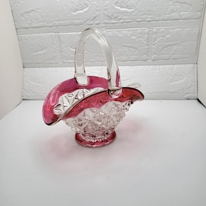 May include: A clear glass basket with a ruby red rim and base. The basket has a clear handle and a diamond-cut pattern on the body. The basket is an elegant decorative piece.